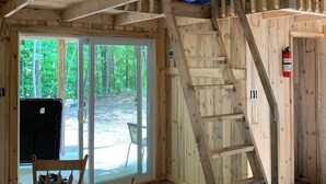 Interior de una cabaña con escalera de madera, zona de comedor con mesa y sillas, y un amplio ventanal corredero que da a un espacio exterior.