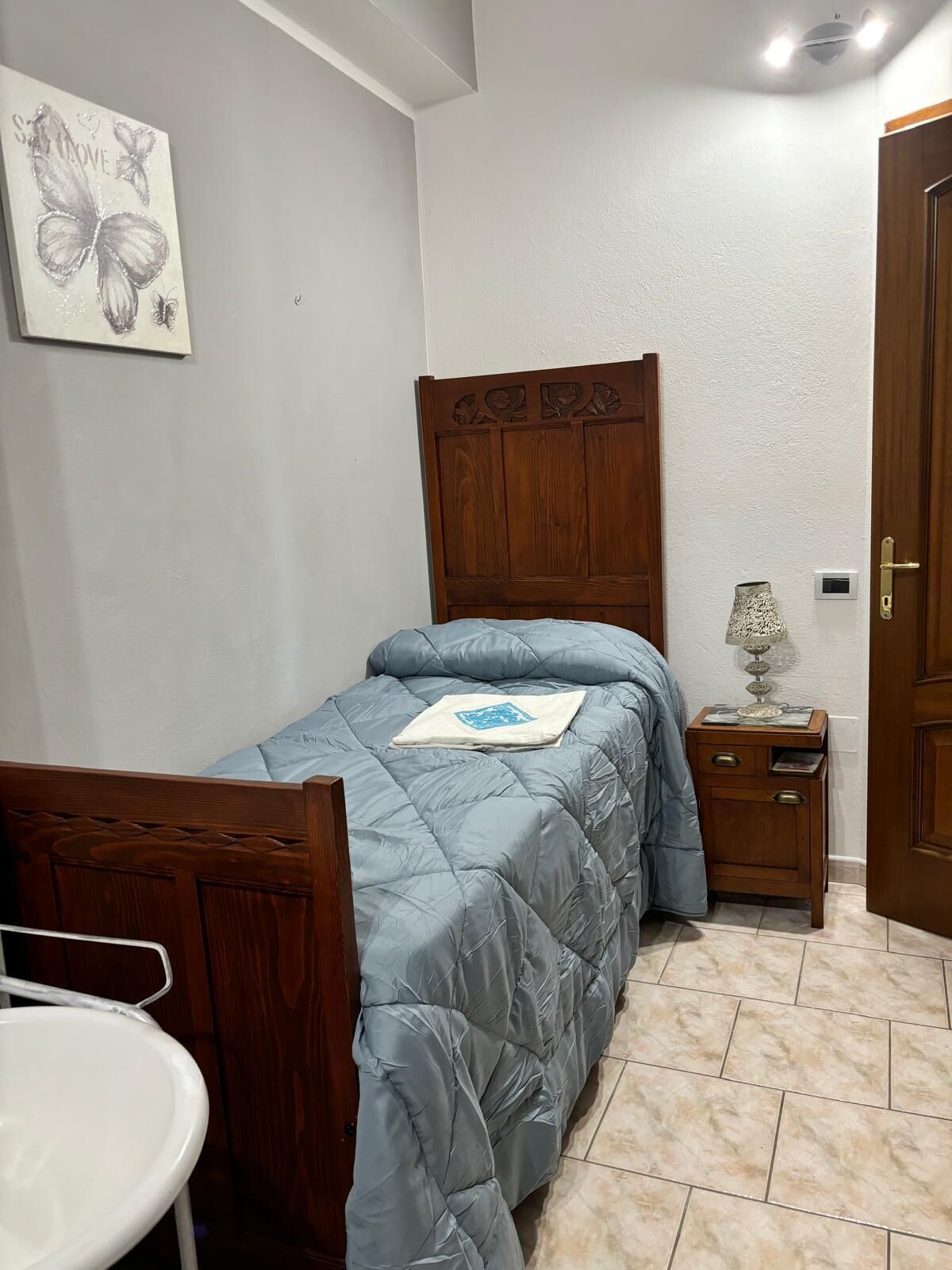 A bedroom with a wooden headboard, a bed with a blue quilt, a bedside table with a lamp, and a white wall with a butterfly artwork.