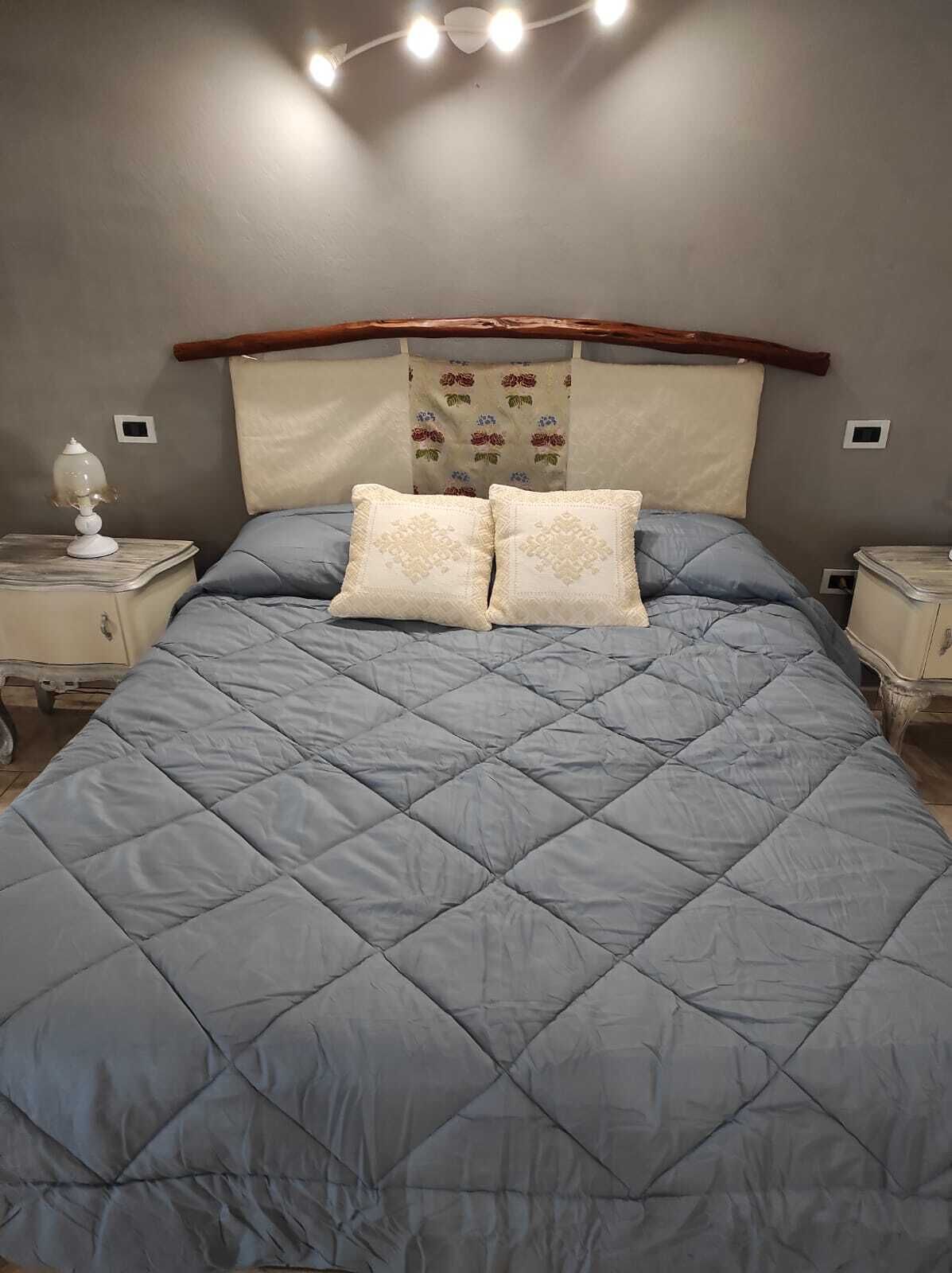 Ein Bett mit einer gesteppten blauen Bettdecke und zwei Kissen, ein hölzernes Kopfteil, Nachttische mit Lampen und eine Wand mit Wandteppich mit Blumenmuster.