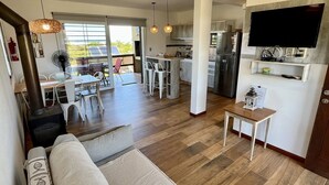 A modern living room with a sofa, a dining area with a table and chairs, a kitchen with a refrigerator and a TV mounted on the wall. - Los Caracoles (Punta del Diablo)