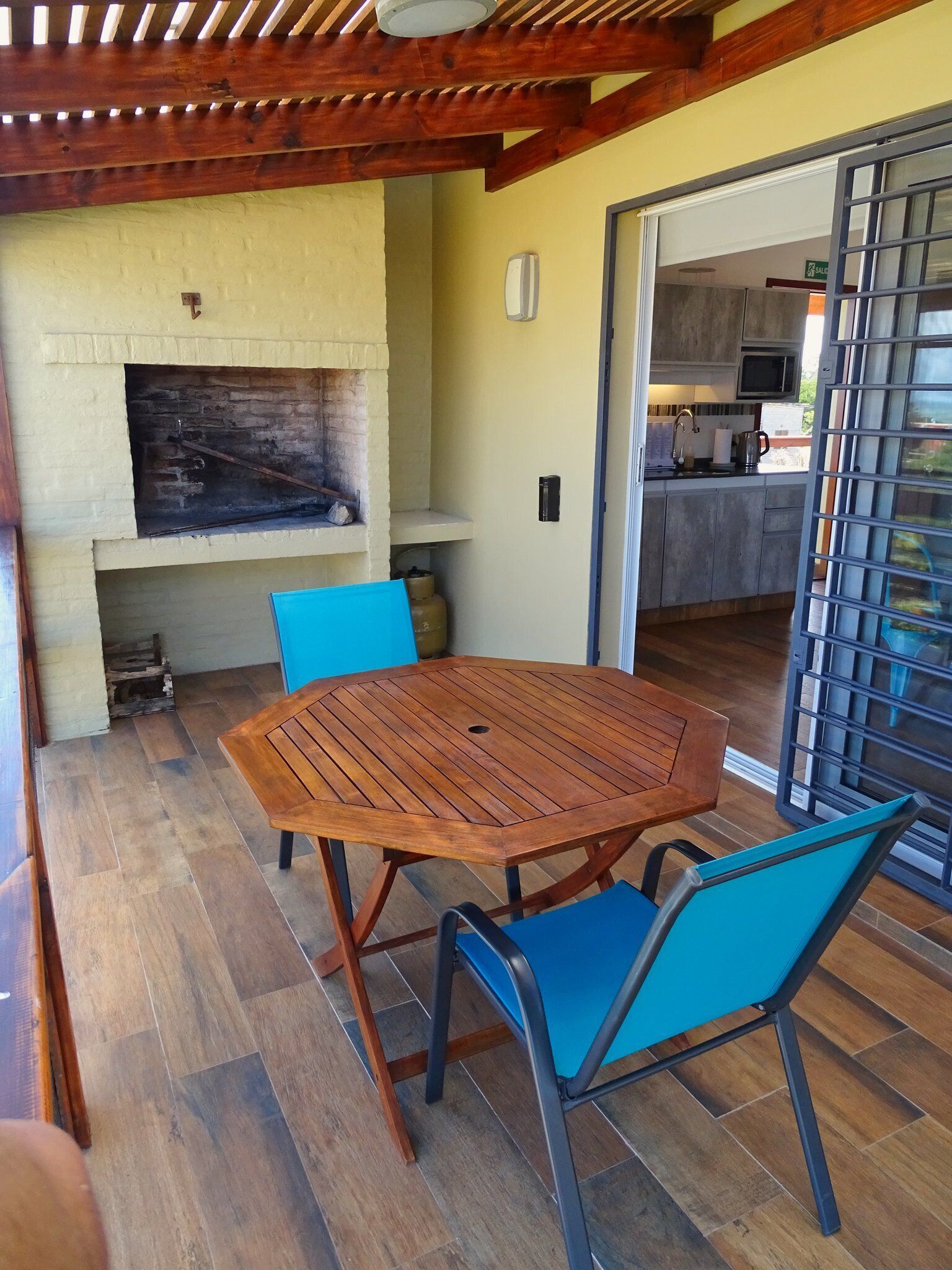 A patio with a wooden table and blue chairs, a brick fireplace, and a kitchen visible through an open door.
