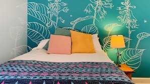 Ein Schlafzimmer mit einer türkisfarbenen Wand mit weißen Pflanzenillustrationen, einer gemusterten Bettdecke und einer Nachttischlampe.
