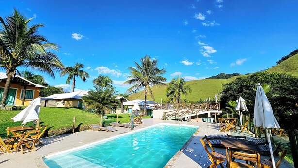 A pool area with wooden loungers and umbrellas, surrounded by palm trees and a hillside.