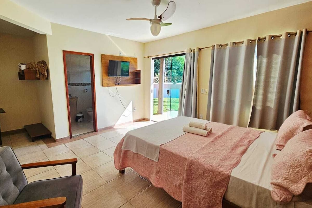 A bedroom with a bed, a chair, a TV, and a bathroom visible through an open door.
