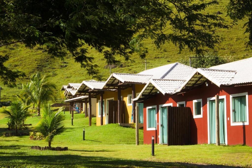 A row of single-story cabins with sloped roofs, each with a door and windows, set against a backdrop of lush green hills and palm trees.