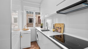 A modern kitchen with white cabinetry, a built-in dishwasher, a washing machine, a stovetop, and a window with a view of a building outside.