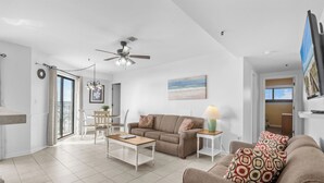 A spacious living room with a sofa, coffee table, and a ceiling fan. There is a dining area with chairs and a table, and a kitchen area with a counter and cabinets.