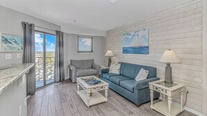 Una sala de estar con un sofá gris, un sofá azul y una mesa de centro. Hay dos lámparas sobre mesitas auxiliares, un cuadro en la pared y una ventana con cortinas.
