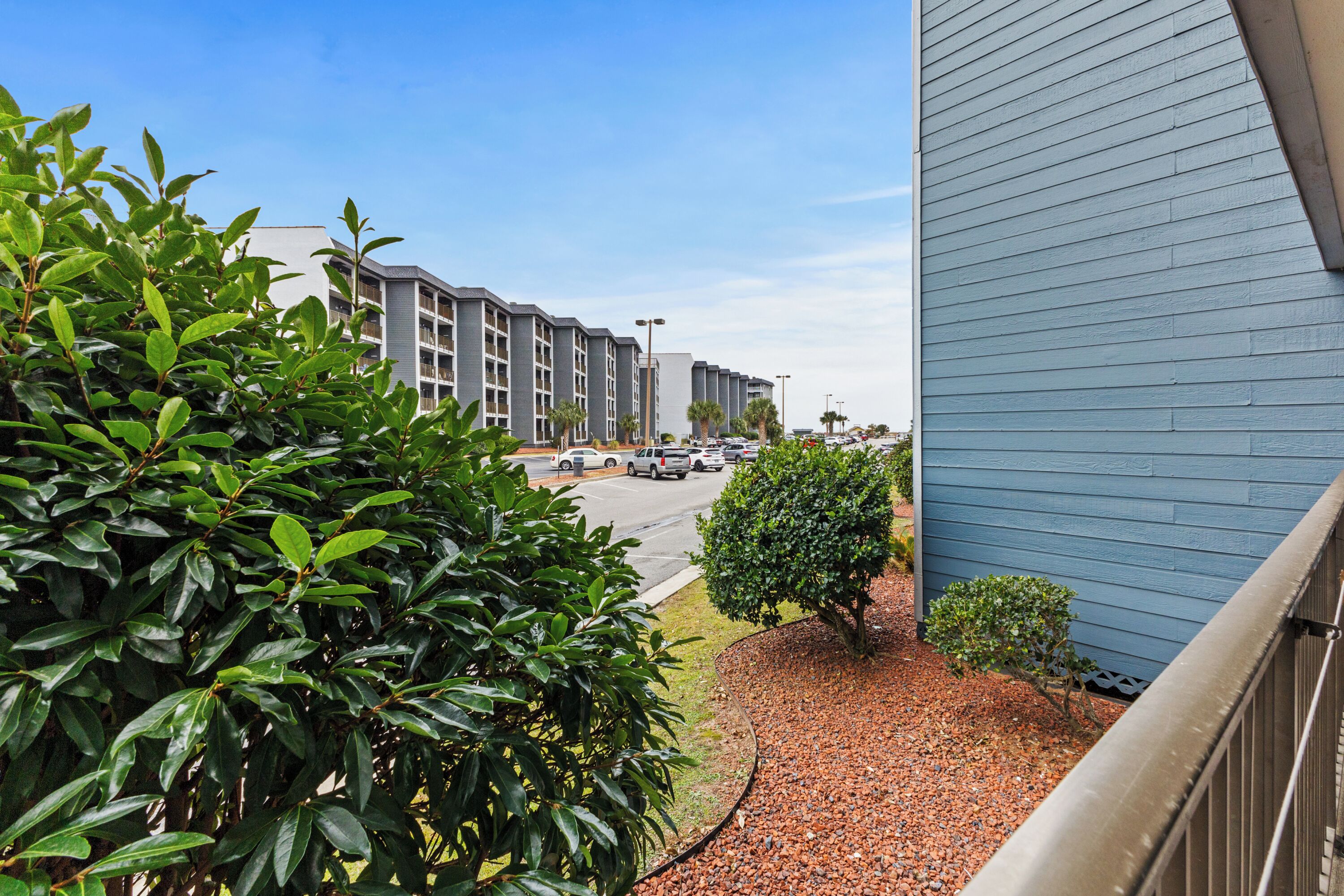 On voit un bâtiment à la façade bleue, un parking avec plusieurs voitures et un petit arbre au premier plan.