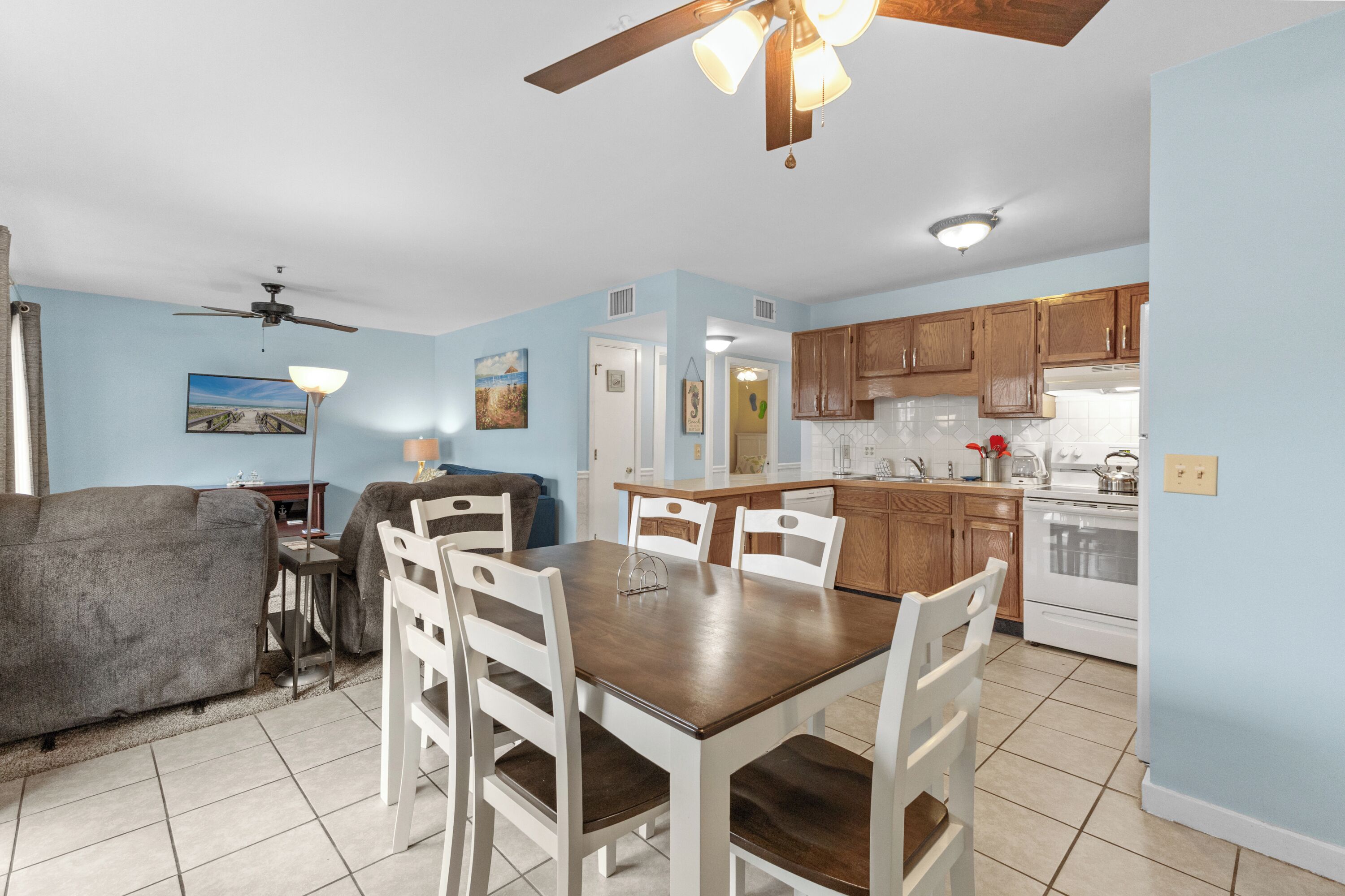 A dining area with a wooden table and white chairs, a ceiling fan, and a kitchen in the background.