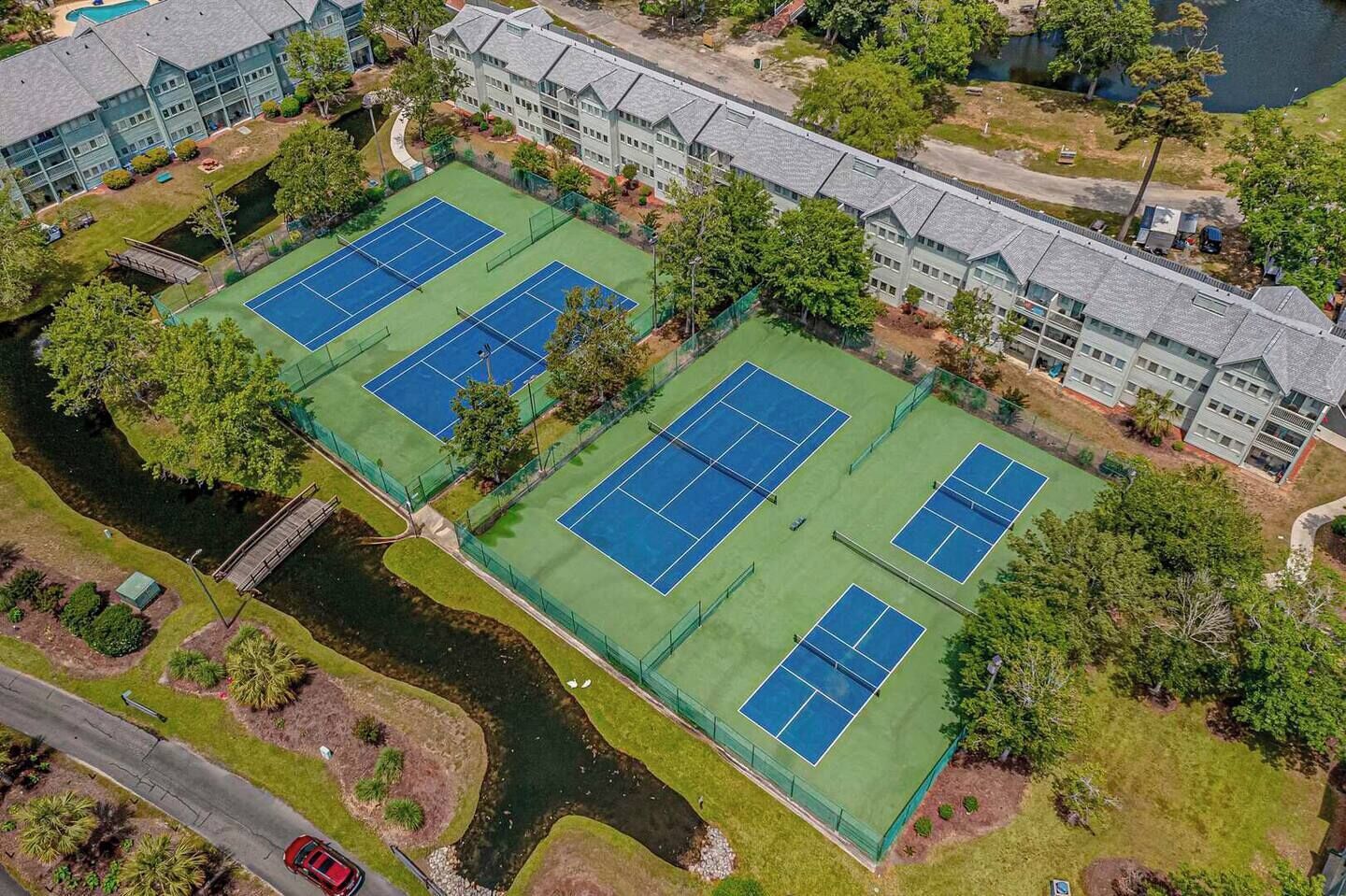 An aerial view of a tennis court complex with multiple courts, surrounded by trees and a building.