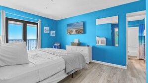 Ein Schlafzimmer mit einem großen Bett, einem Schminktisch, einem Gemälde und Meerblick durch ein Fenster.