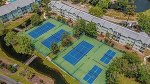 Vista aérea de un complejo de canchas de tenis con varias canchas, rodeado de árboles y un edificio.