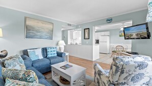 A modern living room with a blue sofa, white coffee table, and patterned armchair. A flat-screen TV is mounted on the wall, and a dining area is visible in the background. - Myrtle Beach Resort A0327 (Myrtle Beach)