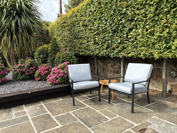 Un espace extérieur aménagé avec deux chaises et une petite table, entouré d’un sol en pierre et d’une végétation luxuriante.