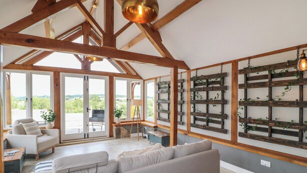 A spacious living area with a high ceiling and wooden beams, a large sofa, and a view through large windows to the outdoors. - New Elms Farmhouse (Derby)
