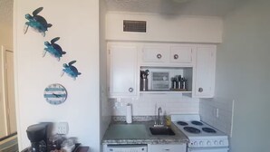 A compact kitchen with white cabinets, a built-in microwave, a stove, and a dishwasher. There are decorative wall art pieces and a clock on the wall. - Private Bayview Condo (Corpus Christi)