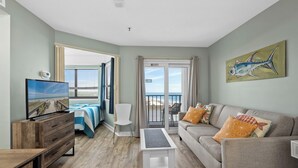 Ett vardagsrum med en tv, en soffa, ett soffbord och utsikt över stranden genom balkongdörrarna.