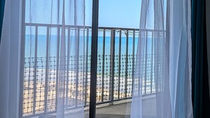 A view through a window with sheer curtains, revealing a balcony with a metal railing and a sandy beach. - 2 Bedroom Ocean View | Myrtle Beach Resort RT1015 (Myrtle Beach)