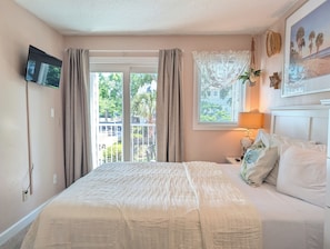 A bedroom with a bed, a TV mounted on the wall, a window with curtains, a bedside table with a lamp, and a framed picture on the wall.