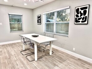 Un espace repas moderne avec une table blanche, des chaises en métal et un bol noir. Il y a deux fenêtres équipées de stores et des œuvres d’art sont accrochées aux murs.