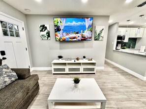A modern living room with a flat-screen TV, a white coffee table, a sofa, and a kitchen area in the background.
