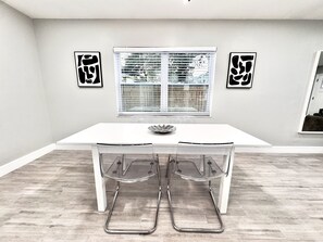 Un espace de salle à manger moderne avec une table et des chaises blanches, une fenêtre avec des stores et des œuvres d’art abstraites accrochées au mur.