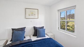 A bedroom with a bed, two pillows, a nightstand, a framed picture on the wall, and a window with a view of greenery.