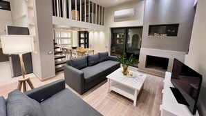 A modern living room with a grey sofa, a coffee table, and a TV. There is a staircase leading to an upper level with a dining area.