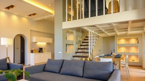 A modern living room with a grey sofa, a coffee table, and a dining area with a table and chairs. There is a staircase leading to an upper level.