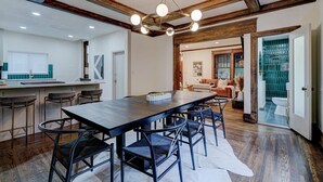 A spacious dining area with a long table and chairs, a kitchen area with bar stools, and a view into a living room with a sofa and a green-tiled bathroom. - Downtown Tulsa’s Emerald on Carson (Tulsa)