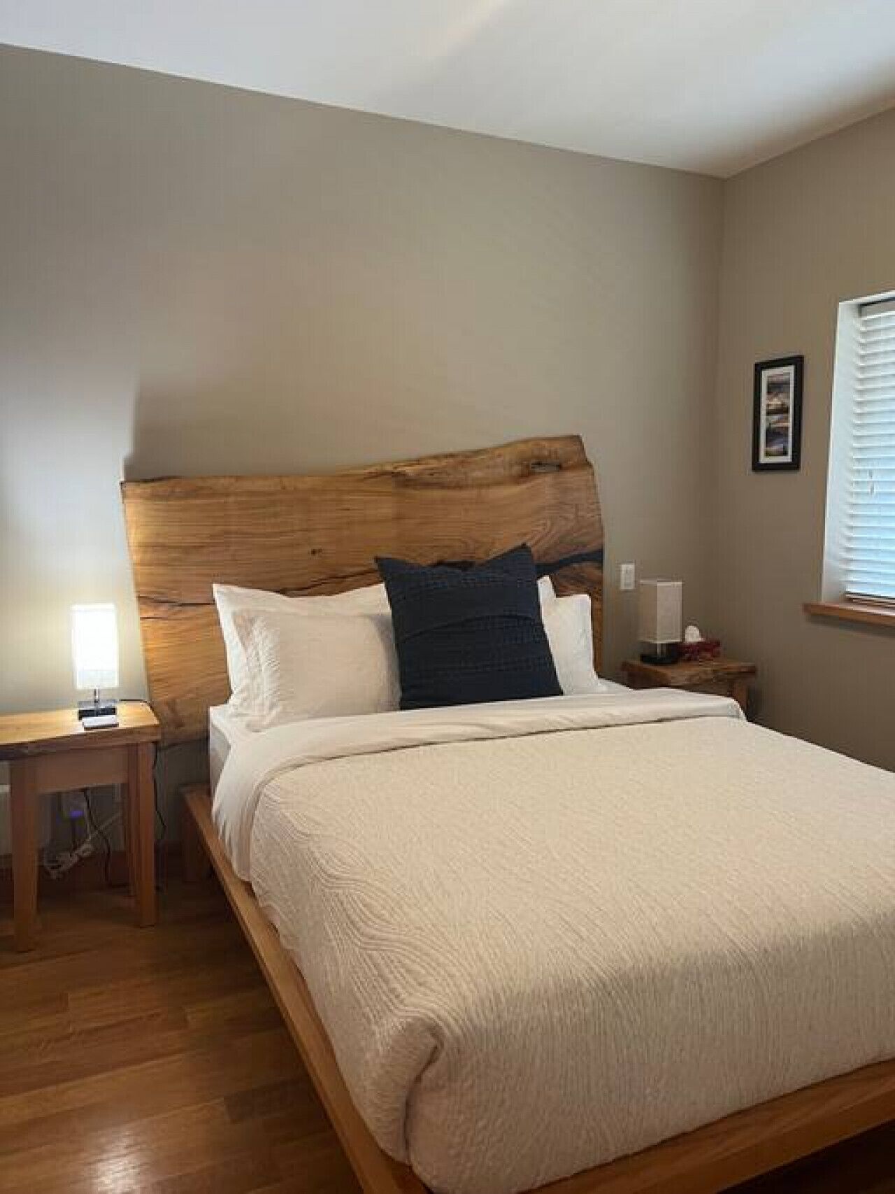 A bedroom with a wooden headboard, a bed with white and dark pillows, a bedside table with a lamp, and a window with blinds.