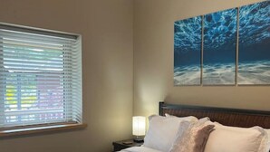 Ein Hotelzimmer mit einem großen Bett, einem Nachttisch mit Lampe, einem Fenster mit Jalousien und einem Wandbild mit Unterwasserlandschaft.