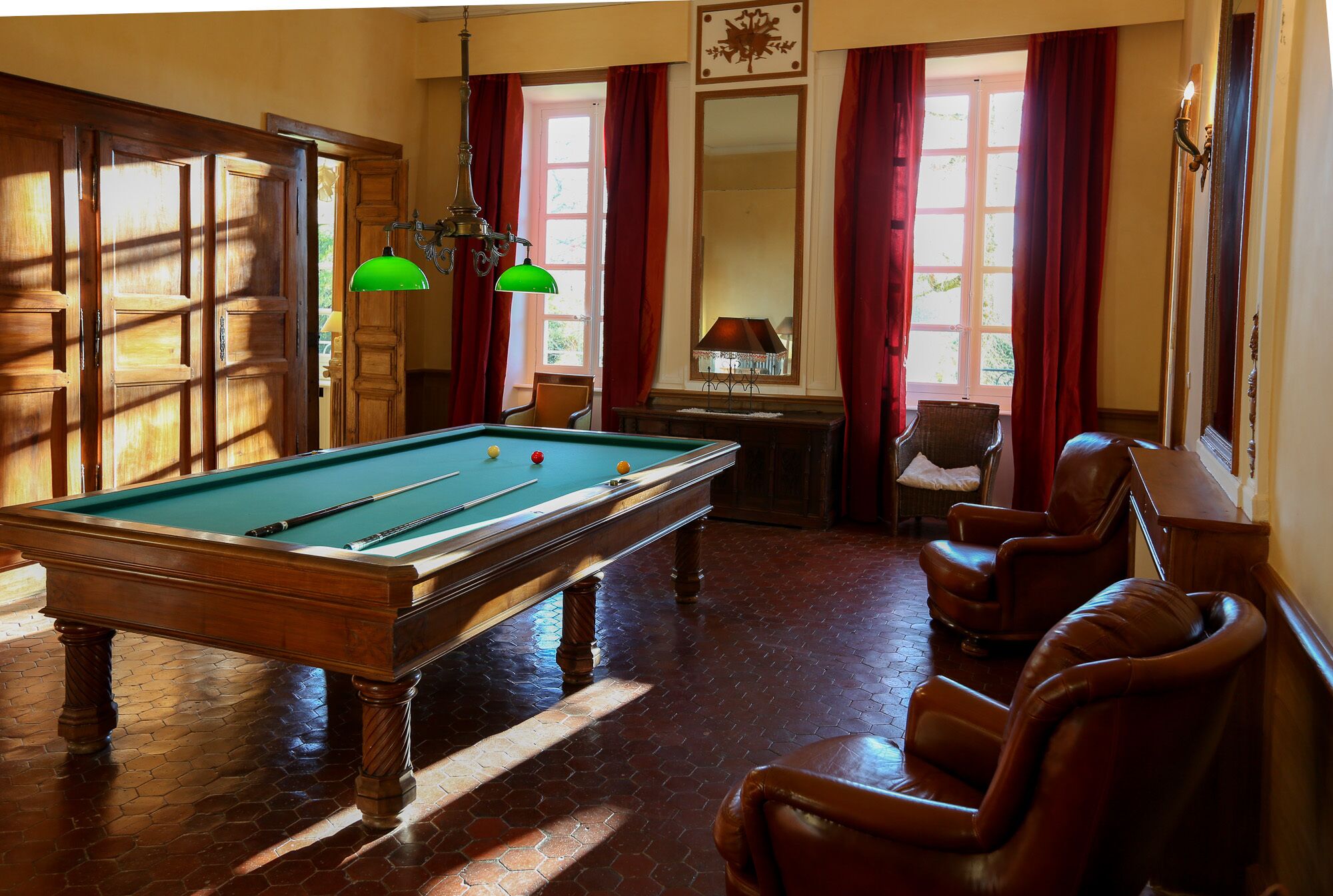 Una mesa de billar en una habitación con suelo de madera, sillones de cuero y una ventana con cortinas rojas.