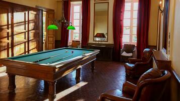 A pool table in a room with wooden flooring, leather chairs, and a window with red curtains.