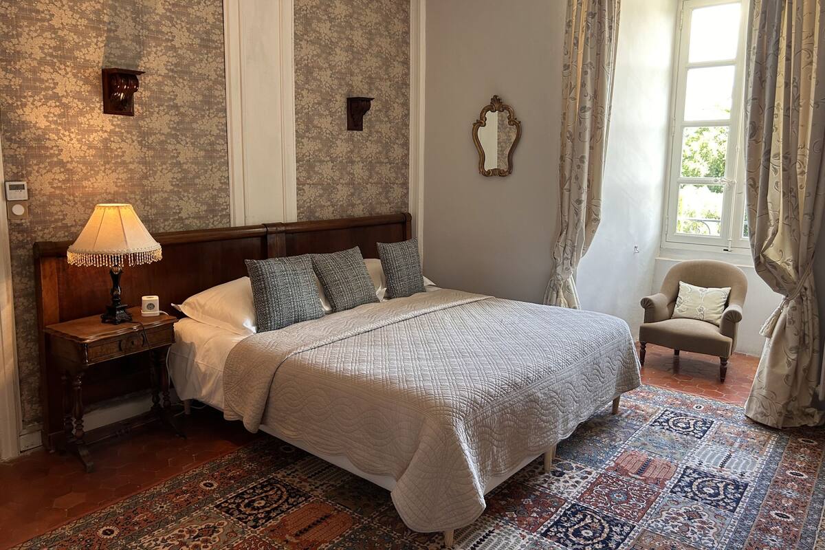 Un dormitorio con una cama grande, una mesita de noche con lámpara, una silla y una ventana con cortinas.