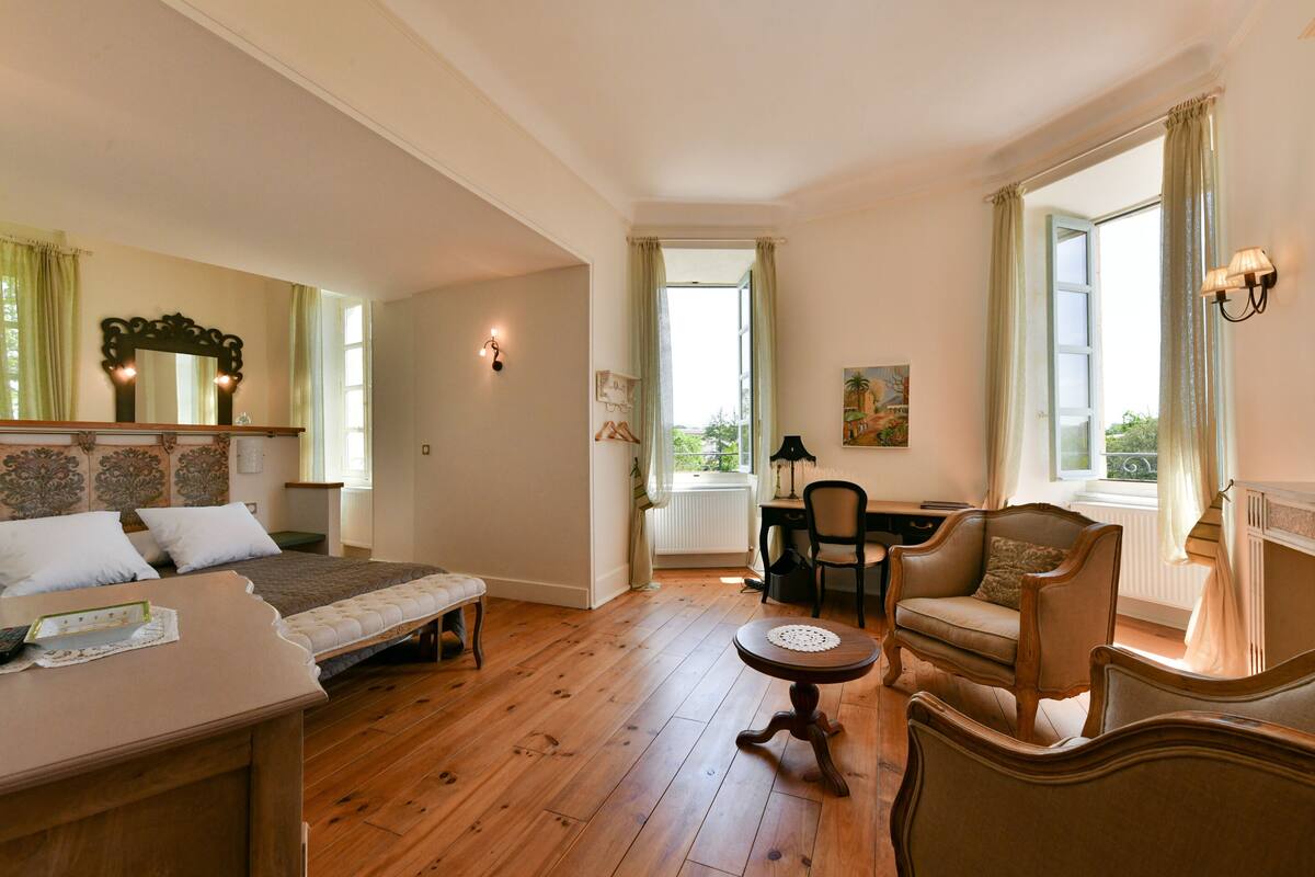 Una habitación amplia con una cama, una mesa de comedor y un sofá. Hay una ventana con cortinas, una lámpara y un cuadro en la pared.