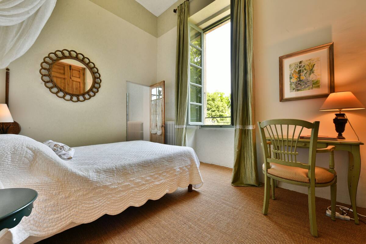 Un dormitorio con cama, una silla, una mesa, ventana con cortinas, un espejo y un cuadro en la pared.