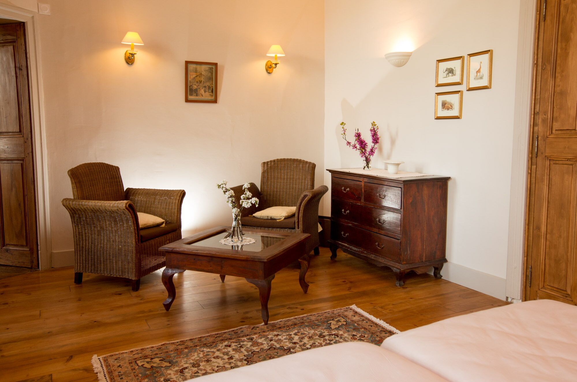 A cozy room with a wooden floor, a bed, two wicker chairs, a wooden dresser, a small table with a vase of flowers, and wall-mounted lamps.