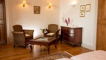 A cozy room with a wooden floor, a bed, two wicker chairs, a wooden dresser, a small table with a vase of flowers, and wall-mounted lamps.