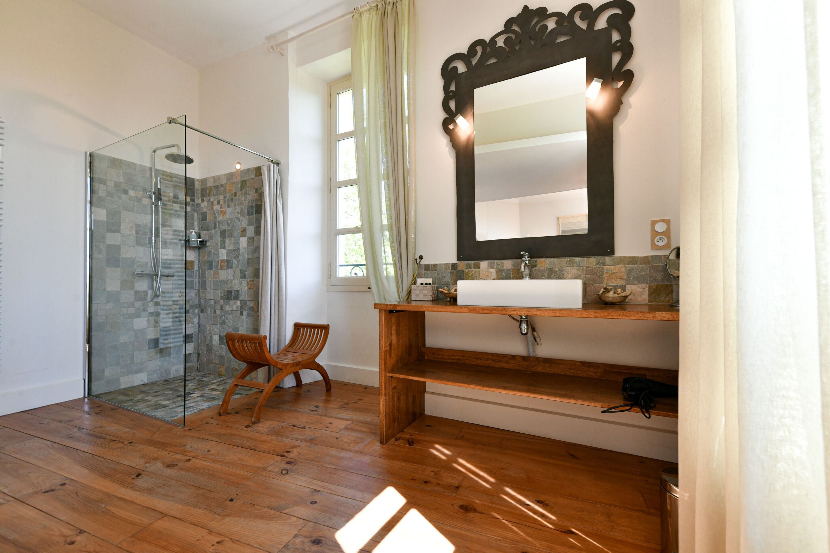 Un baño con ducha de vidrio, suelo de madera, una silla de madera, lavabo con espejo y una ventana con cortinas.