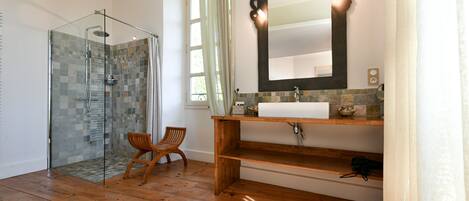 Un baño con ducha de vidrio, suelo de madera, una silla de madera, lavabo con espejo y una ventana con cortinas.