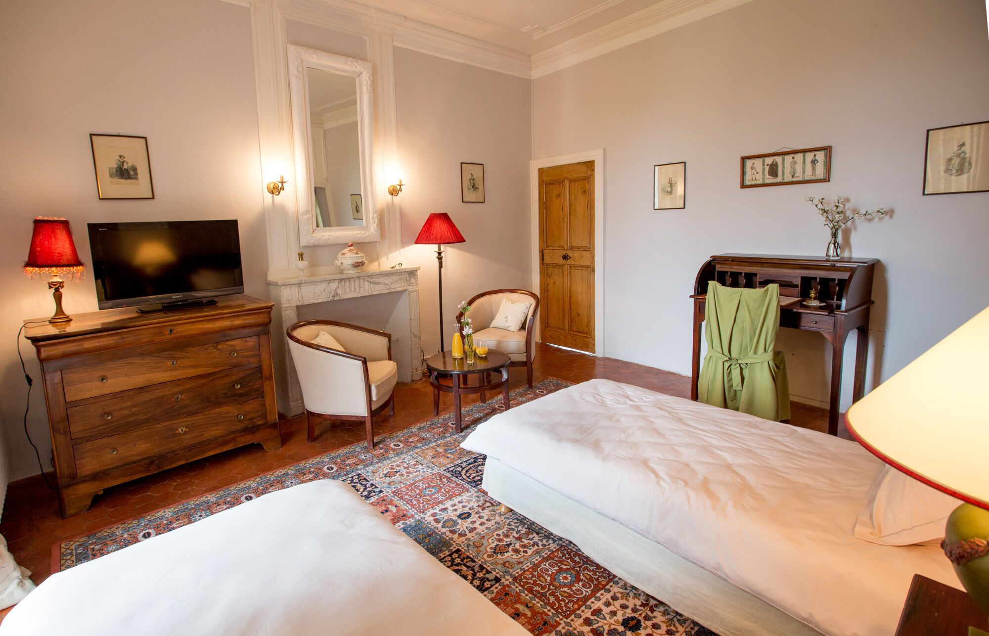 A hotel room with two beds, a desk with a television, a chair, a wooden wardrobe, and a desk lamp.