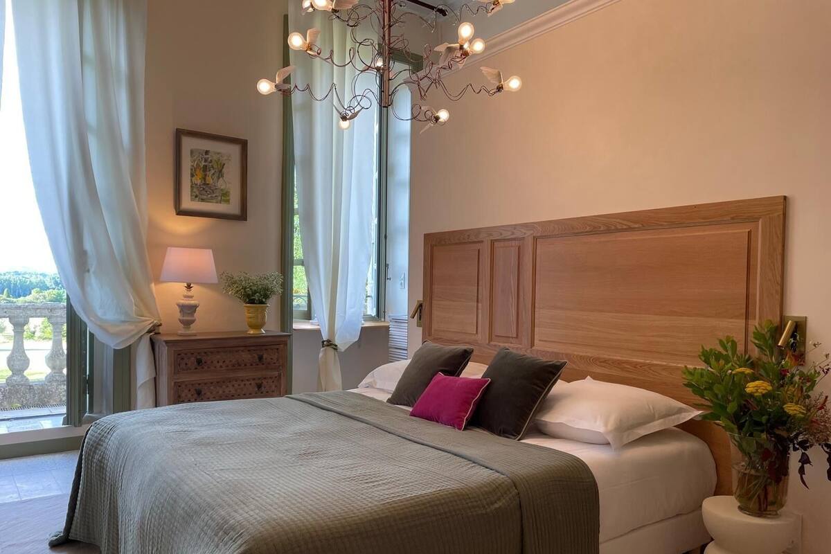 Un dormitorio con una cama grande, un cabecero de madera, una araña, una mesita de noche con lámpara y una ventana con cortinas.