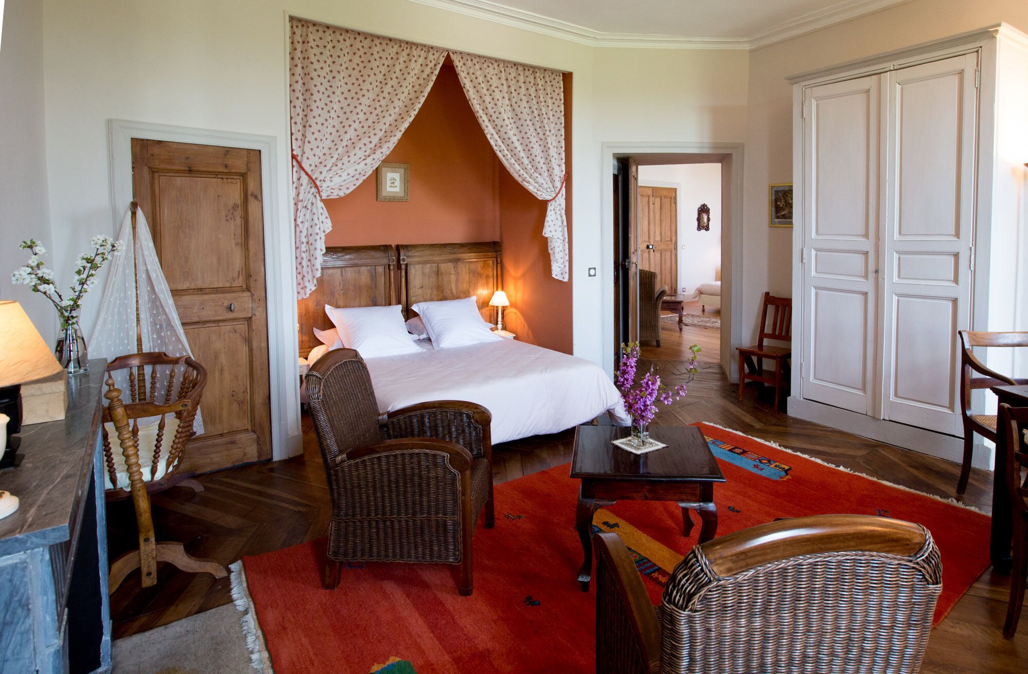 Une chambre d’hôtel avec un lit, deux fauteuils en osier, une table, un tapis rouge et une armoire en bois.