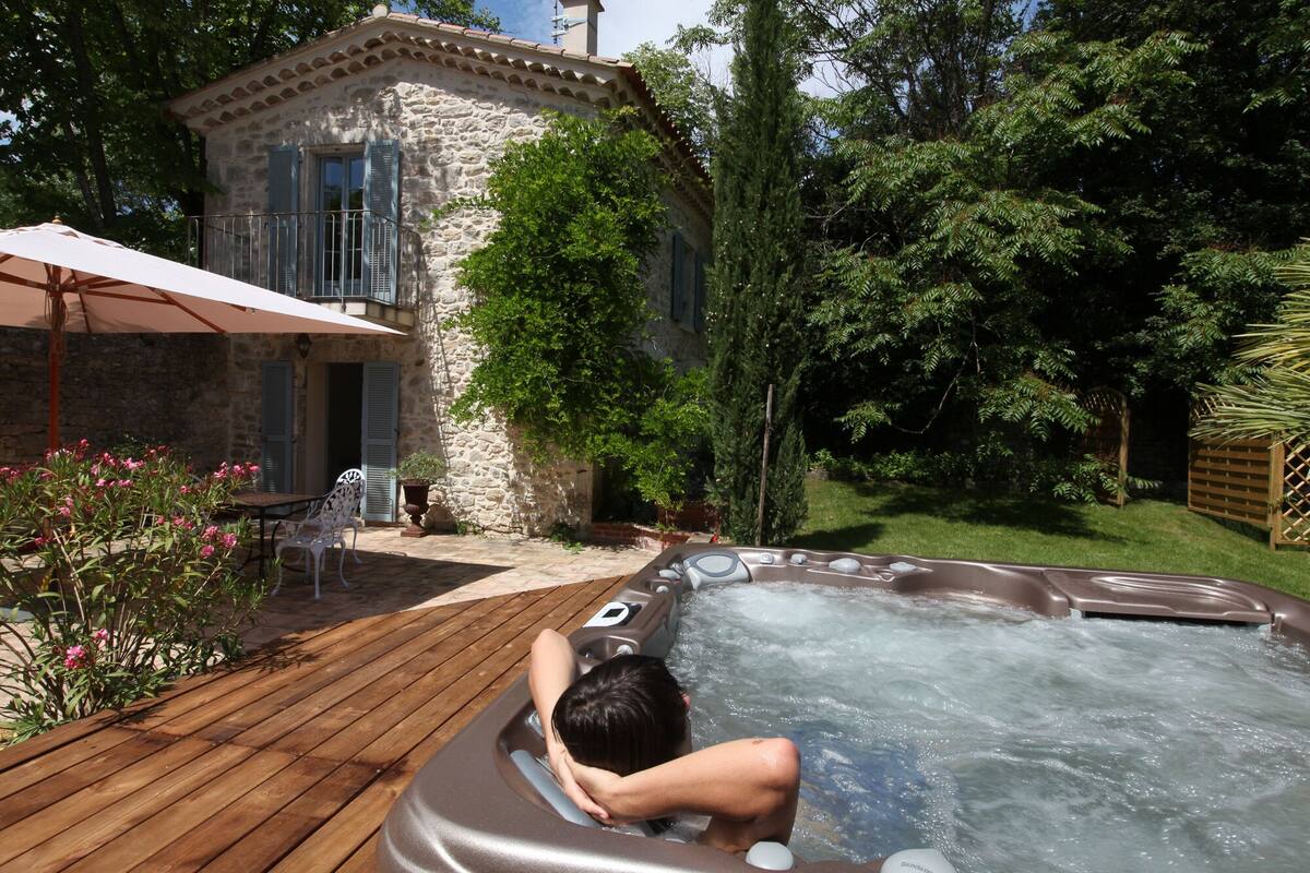 Un hombre relajándose en un jacuzzi al aire libre, con un edificio de piedra y exuberante vegetación de fondo.