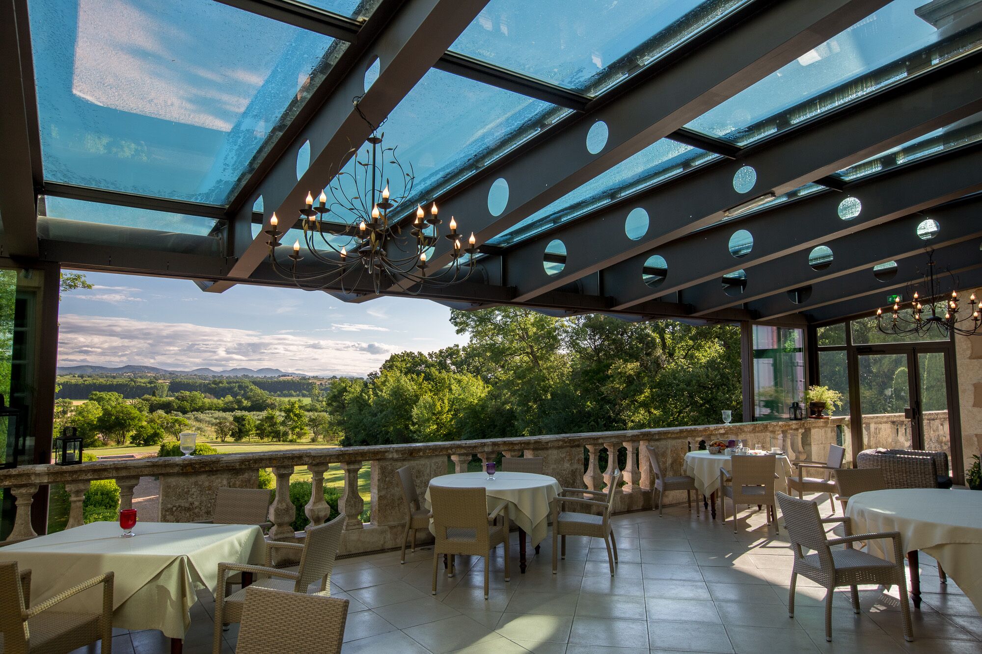 A covered outdoor dining area with tables and chairs, a glass ceiling, and a view of the landscape.