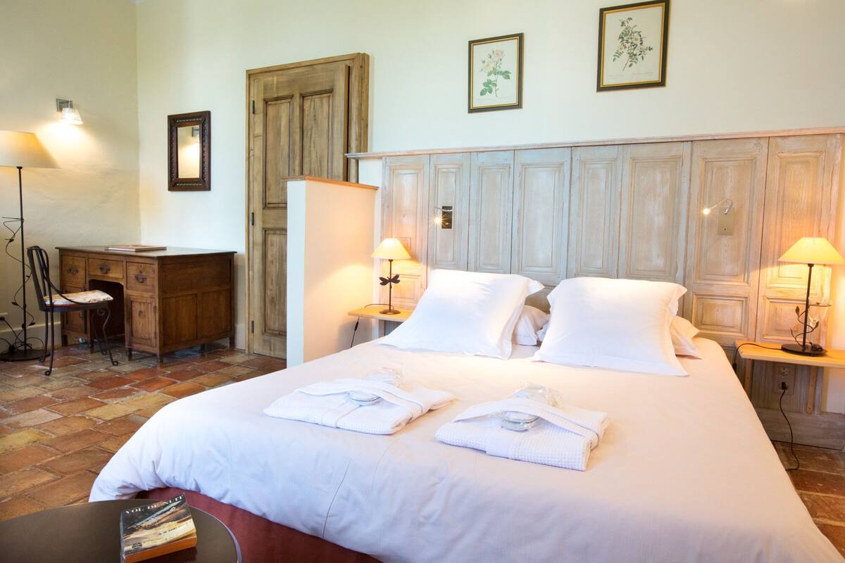 Una habitación de hotel con una cama grande, dos mesitas de noche con lámparas, un escritorio de madera, una silla y una puerta.