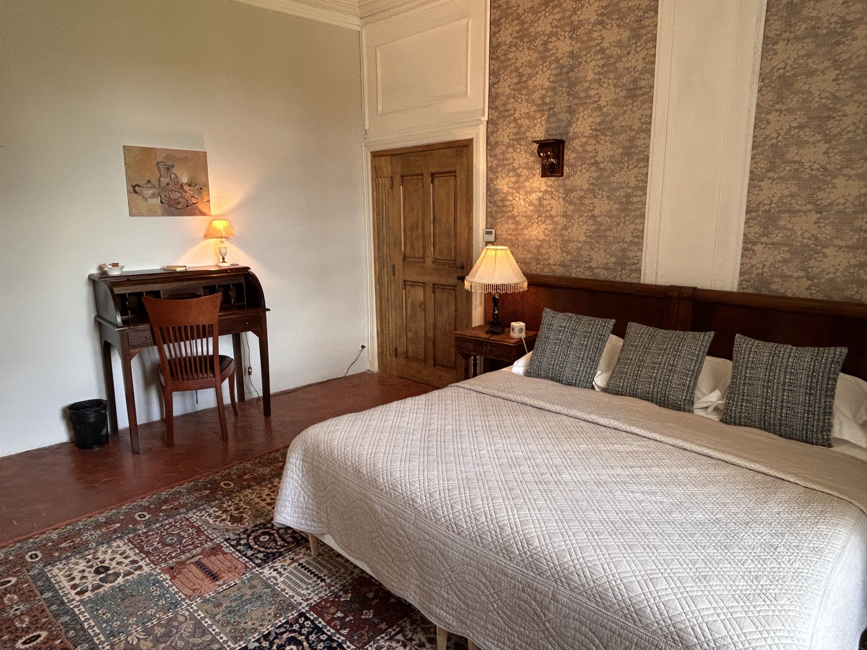 Una habitación de hotel con cama, escritorio, silla, lámparas y un tapete estampado.
