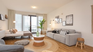 Un salón moderno con un sofá gris, un sillón blanco, una mesa de centro de mimbre y un taburete pequeño. Hay una ventana grande con cortinas, una planta en maceta y cuadros en la pared.